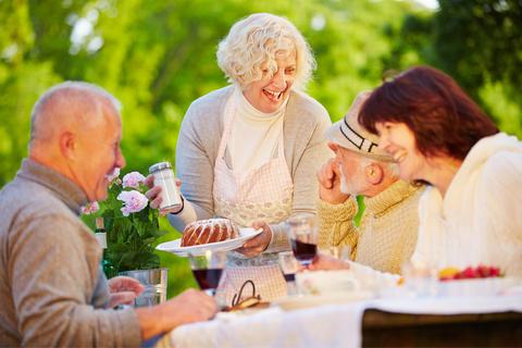 Older men and women enjoying dinner outside.  One older woman holding a cake in one hand and powdered sugar in the other.