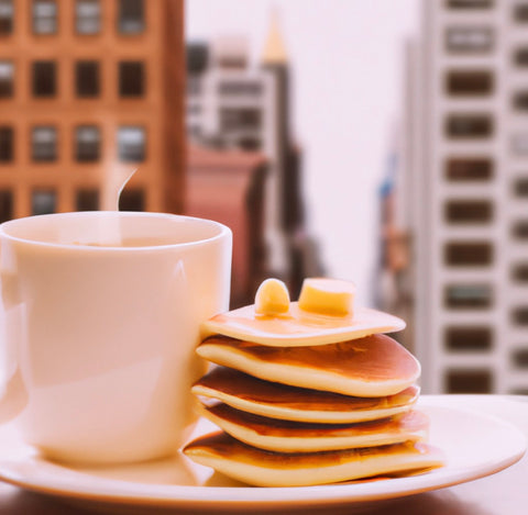 a coffee cup next to a plate of breakfast food such as pancakes or pastries, with a background of a coffee shop or a cozy kitchen setting Picasso Style