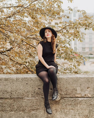 A modern witch sits atop an old wall above the River Seine in Paris, wearing the little black T-Neck Shift Dress cut from sustainable organic cotton bamboo viscose