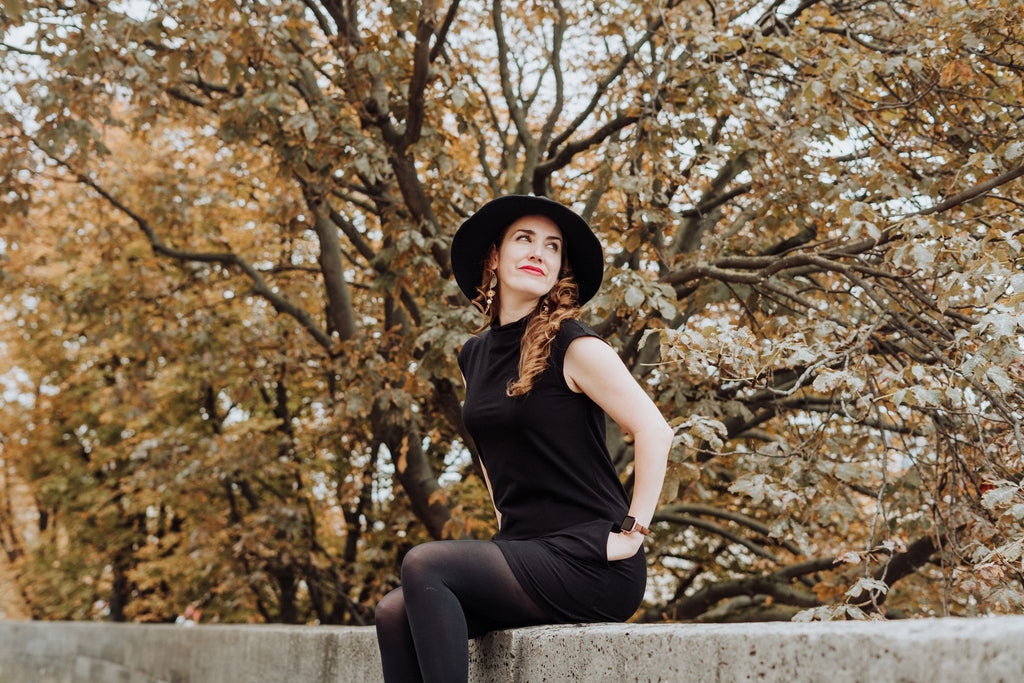 A modern witch sits on an old stone wall along the banks of the Seine River in Paris, wearing black "Witch" hat and DeLaunay organic cotton/bamboo viscose T-Neck Cap Sleeve Dress in black.