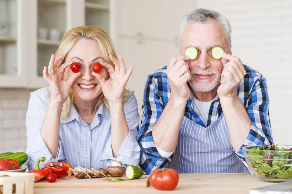 smiling senior couple holding cherry tomatoes and cucumber slices in front of their eyes