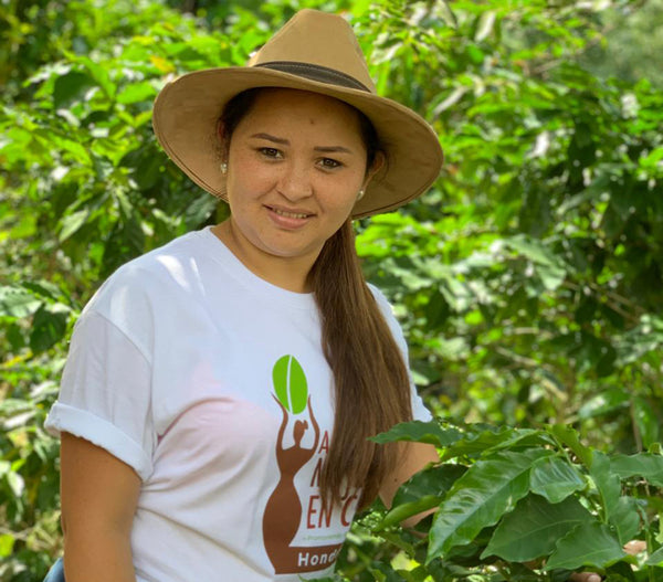 Woman in tan hat smiling with coffee plants behind her.