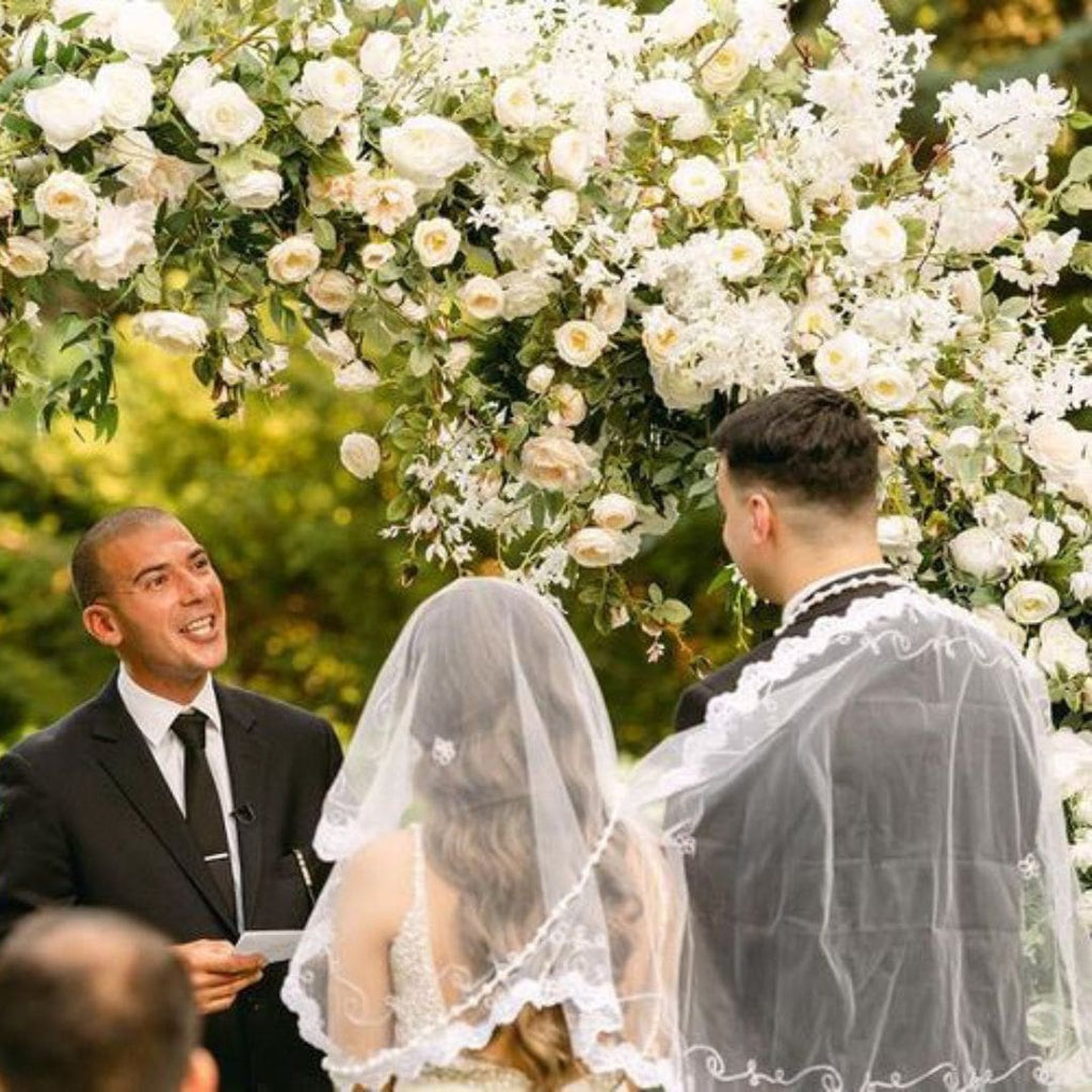 A couple during their unity cord & veil ceremony, standing underneath an arch of flowers, including star jasmine which resembles sampaguita.