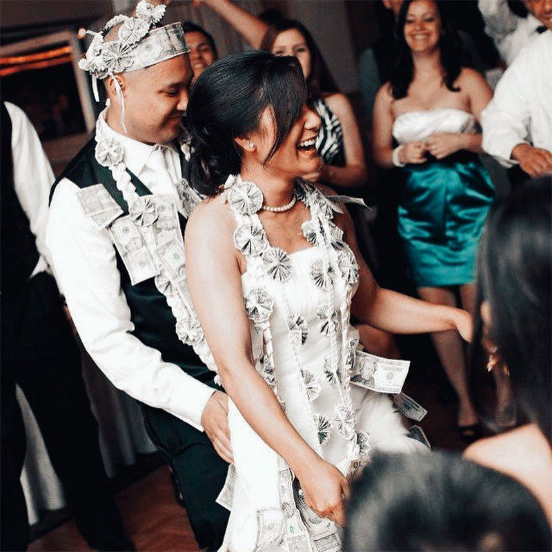 Filipino newlyweds in the middle of their money dance, looking festive in a crown, sash, and garlands made of cash.
