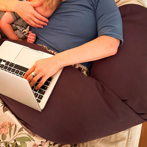 Die Mama arbeitet von zu Hause aus im Homeoffice. Sie ist selbständig und arbeitet am Laptop für ihr Unternehmen. Die Arme liegen entspannt auf dem Stillkissen, welches Sie sich als Laptopunterlage drapiert hat. So kann sie stillen und arbeiten.