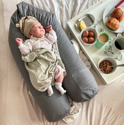 Das Baby liegt auf einem Bio-Stillkissen von Zellmops, während die Mama und der Papa im Wochenbett im Bett frühstücken. 