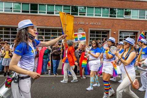 Courtney Danley directing Portland Samba in Portland Pride parade 2022. Blue and white t-shirs and white pants, white fedora with blue band. Portland Fire and Rescue brick building in the backgroung.