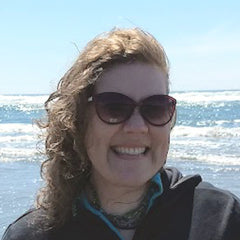 Dannye Chase, writer, standing by the ocean on a bright day, her brown hair windblown, her pale face adorned with sunglasses