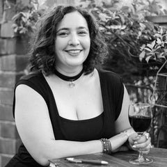 A smiling black and white photo of Tamara M Bailey, a dark-haired, white woman. She sits outside in a garden, smiling and holding a glass of wine