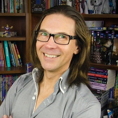Author George Ivanoff in a book-fille study, he has long brown hair, pale skin, wears a button down grey shirt while smiling at the camera