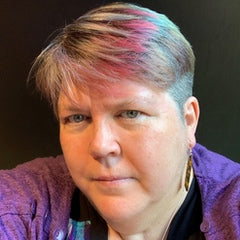 A closeup of writer Narrelle M Harris, her white face is somewhat serious, her short hair is silvery and highlighted with purple-pink accents, and she wears a purple shirt