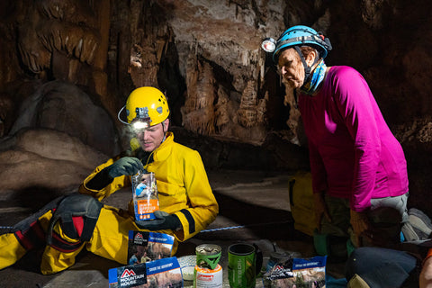 Phyliss overlooks shoulder of man in cave stirring food in Mountain House pouch