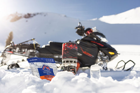 Mountain House Yellow Curry pouch with camp stove in snow with snowmobile behind it.