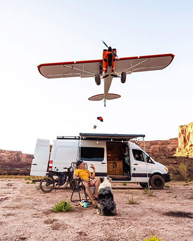 Plane air dropping Mountain House food to Jerimiah who is sitting in camp chair in front of camper van and dirt bike