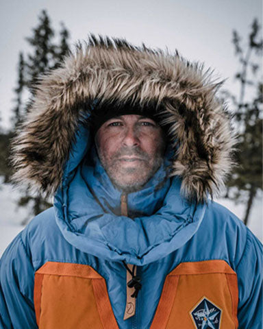 2022 Mountain House Ambassador Jeremiah Young in blue and orange snow coat with fur-lined hood on his head, standing in snow covered forest.