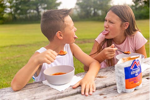 Two kids making faces at one another while they eat Mountain House Spaghetti at a picnic table.