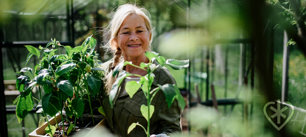 An elderly woman smiling in her garden, in front of a greenhouse, she's holding planters that have sprouted plants