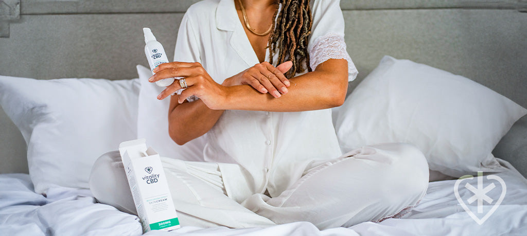 Woman sitting cross-legged on bed, applying Vitality CBD Skin Cream to her forearm