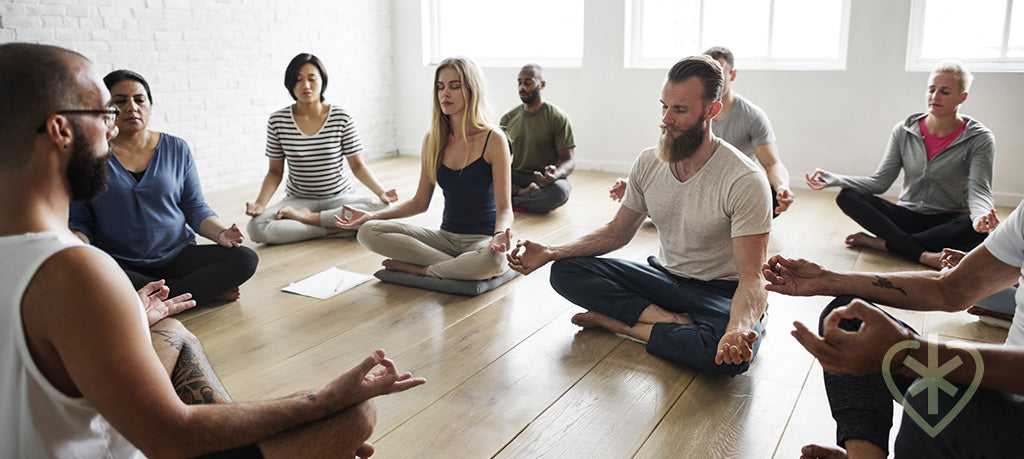 A yoga class with several students facing their teacher, all in lotus pose, breathing deeply