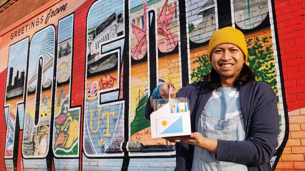 Egads co-owner Remy Darby smiles in front of a colorful mural as he holds a set of Toledo Spark drinkware.