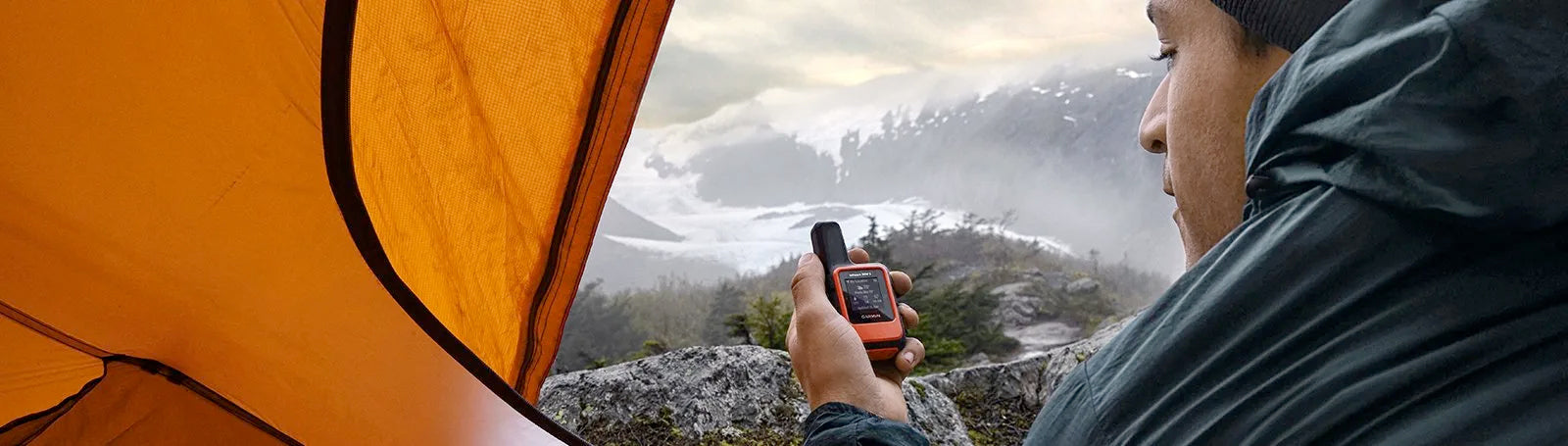 Man in a tent using Garmin InReach Mini 2 while looking out over mountains