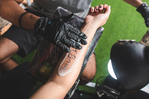 A tattooist cleans a fresh name tattoo on a client's forearm with gentle soap