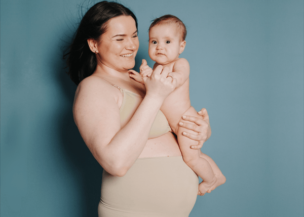 Mum holding her new born baby and smiling, embracing her postpartum body after birth.