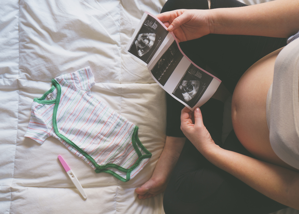 Pregnant mother looking at her ultrasound of her new baby on a bed. There are baby clothes in front of her and she is showing her pregnant belly.