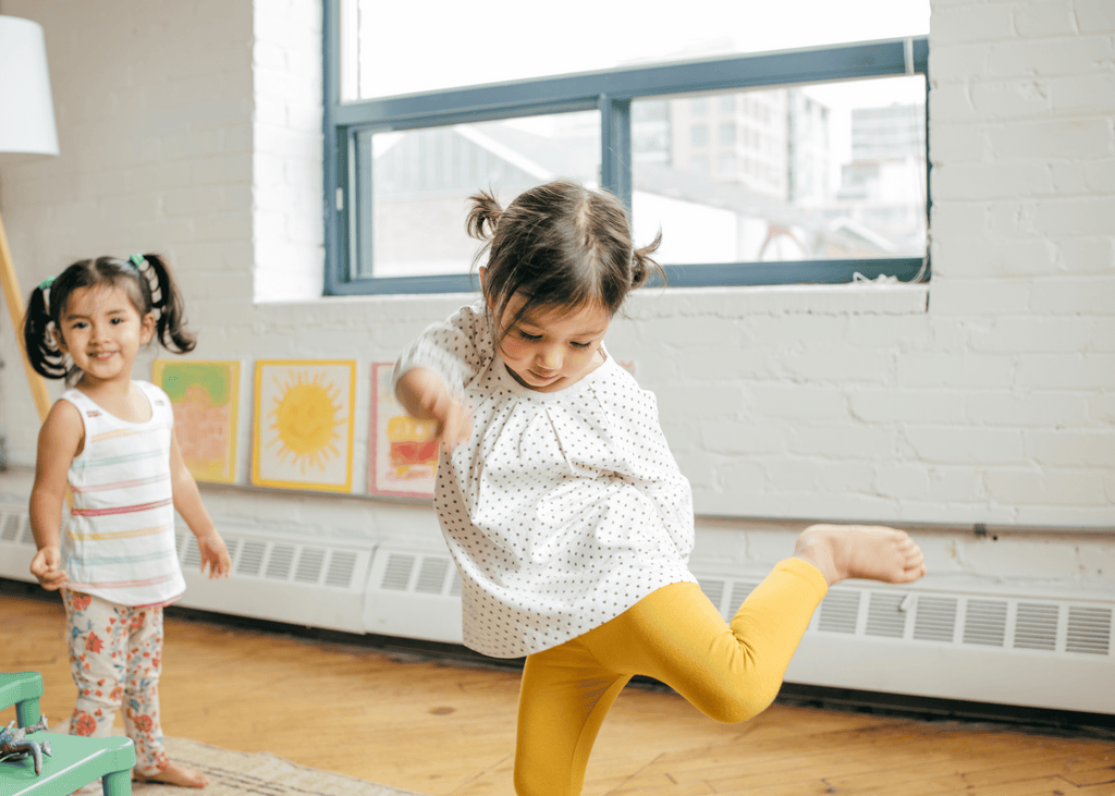 A young toddler playing with her friend at school, dancing happily with one leg in the air.