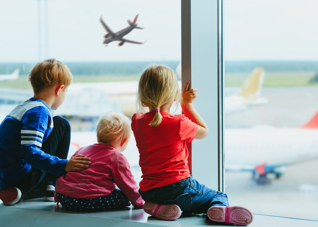 3 small children waiting to catch a flight at an airport and watching planes from the waiting area