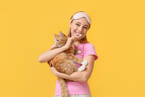 Woman in pink pajamas holding orange tabby cat with toothbrush in her mouth. Yellow background.