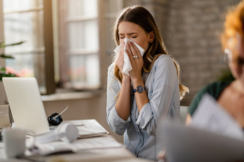 Young woman at her desk sneezing and holding a tissue to her nose.