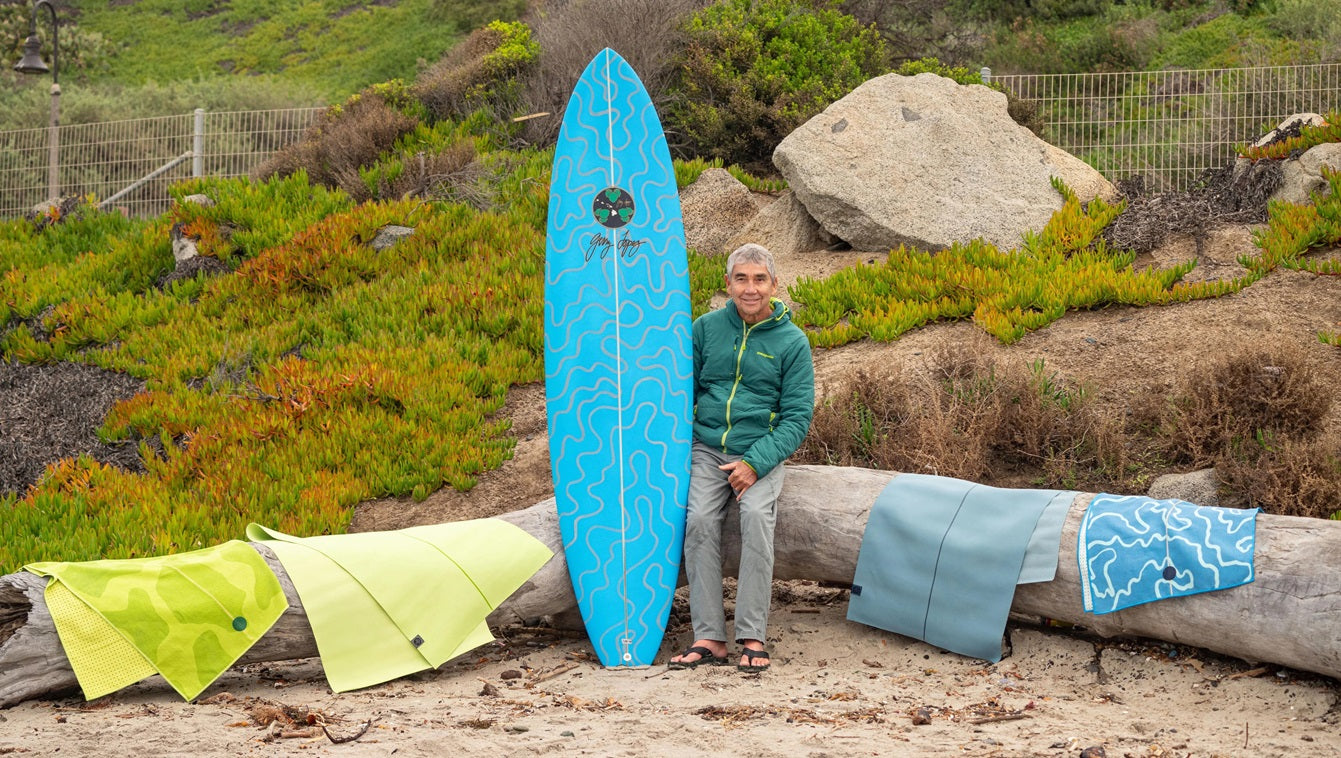 Gerry Lopez am Strand, auf einem Baumstamm sitzend, mit einem blauen Surfbrett und Manduka-Produkten