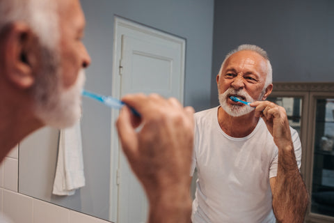 A mature man brushes his teeth in his bathroom, with his reflection in the mirror visible.