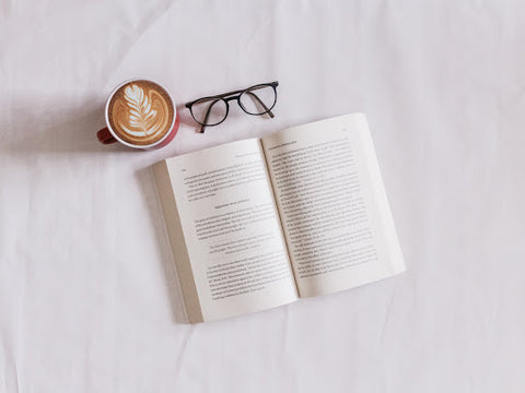 An open book, a pair of reading glasses and a latte on a bed spread, for Ivy Leaf Skincare blog
