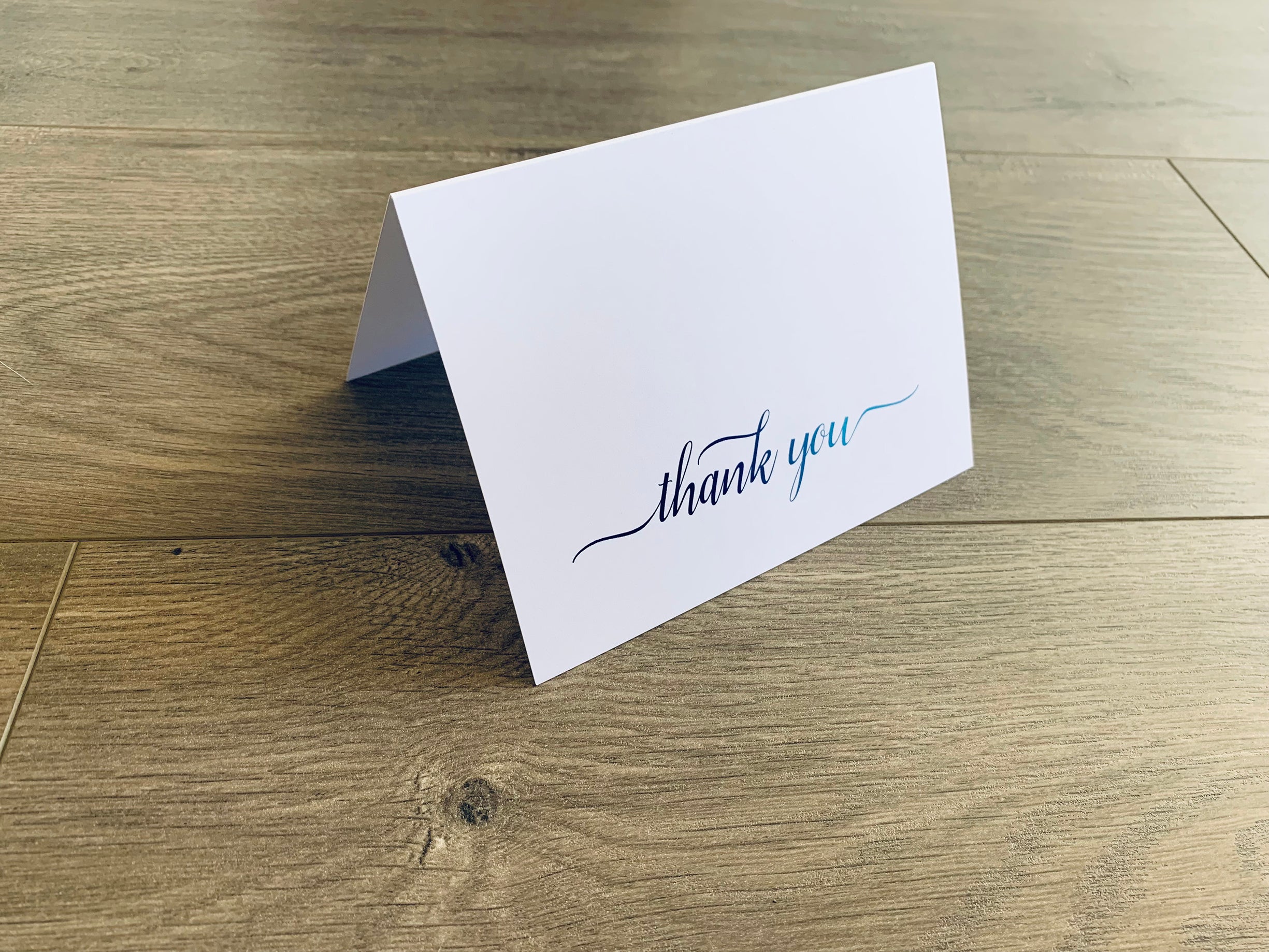A white notecard with a script font that reads "thank you" sits on a wooden floor. The font fades from navy to light blue.