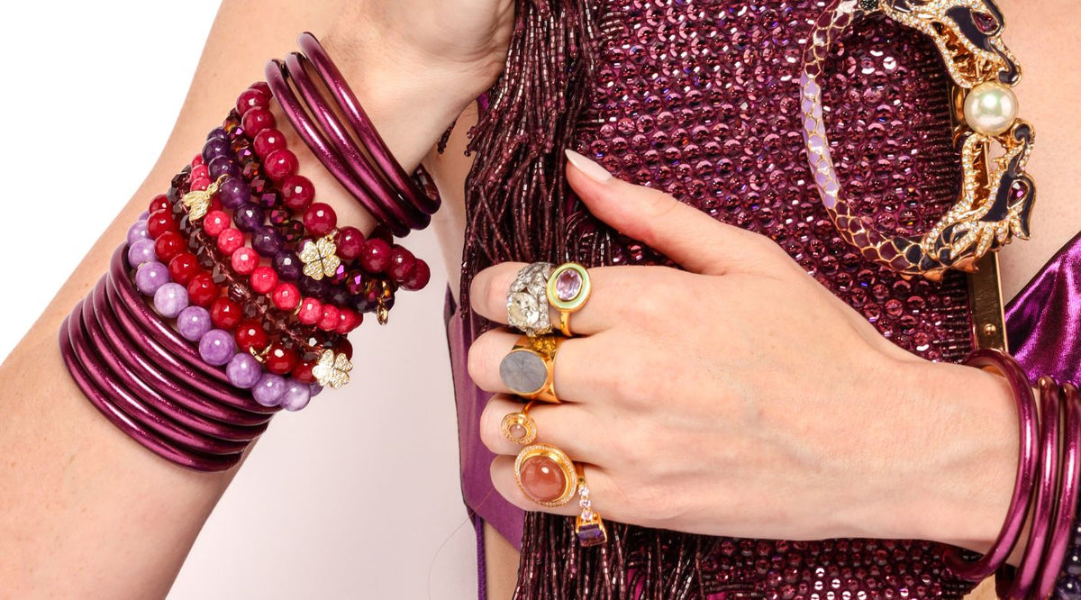 Woman holding purple jeweled purse wearing BuDhaGirl Amethyst All Weather Bangles, Amethyst Chamonix 5 Bracelet set, Aubergine Chamonix 5 bracelet set, Starburst Ring, Pink Camellia Ring,  Purple Amethyst Princess Ring, Peach Moonstone Maxi Orbit Ring, Starburst Ring, Labradorite Starburst Ring, Gold Canasta Ring, Golden Rutile Diva Ring | BuDhaBrief by BuDhaGirl