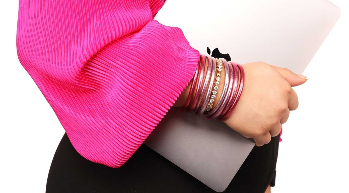 Woman in pink shirt and black slacks wearing BuDhaGirl Carousel Pink All Weather Bangle Bracelets and Carousel Pink Etoile bangle in clear while holding Apple laptop computer | BuDhaBrief by BuDhaGirl