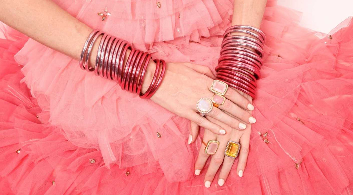 Woman wearing pink dress with Carousel Pink All Weather Bangle bracelets on both arms wearing BuDhaGirl Carousel Pink Nebula Rings, Nebula Ring in Rainbow Moonstone, Nebula Ring in Pink and Green, Nebula Ring in Citrine Amethyst and Camellia Ring in Pink Chalcedony on hands | BuDhaBrief by BuDhaGirl