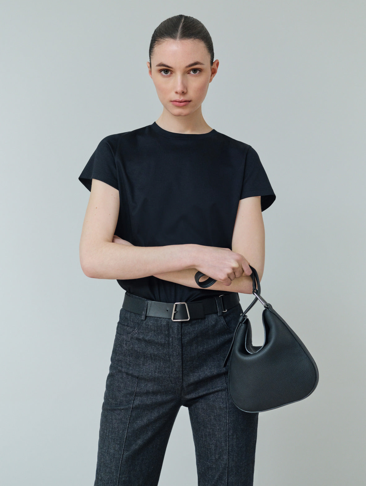 Ein Model posiert in einem dunklen Essentials T-Shirt, einer Denim-Hose und Tasche von Akris