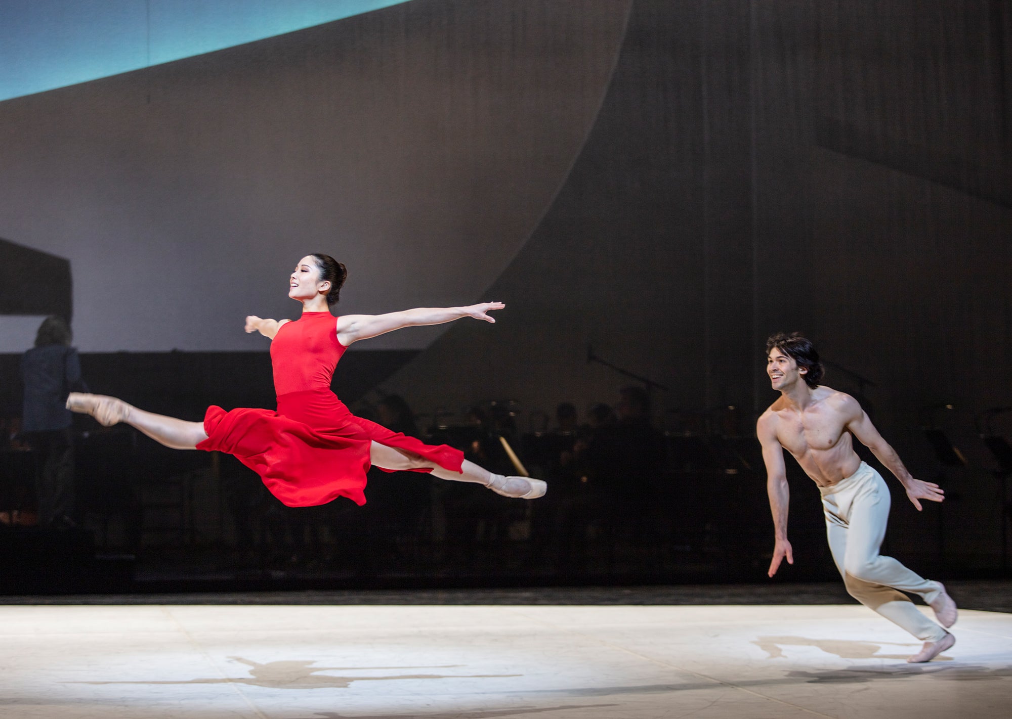 Ballettänzerin in rotem Akris Kleid springt und macht gleichzeitig einen Spagat während ein Ballettänzer ihr nachtanzt