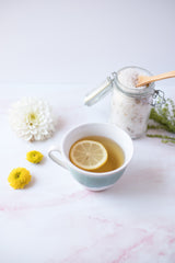 A cup of tea with a slice of lemon in front of a jar of body scrub. 
