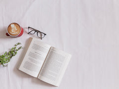 An open book laid out beside a pair of reading glasses and a coffee. 