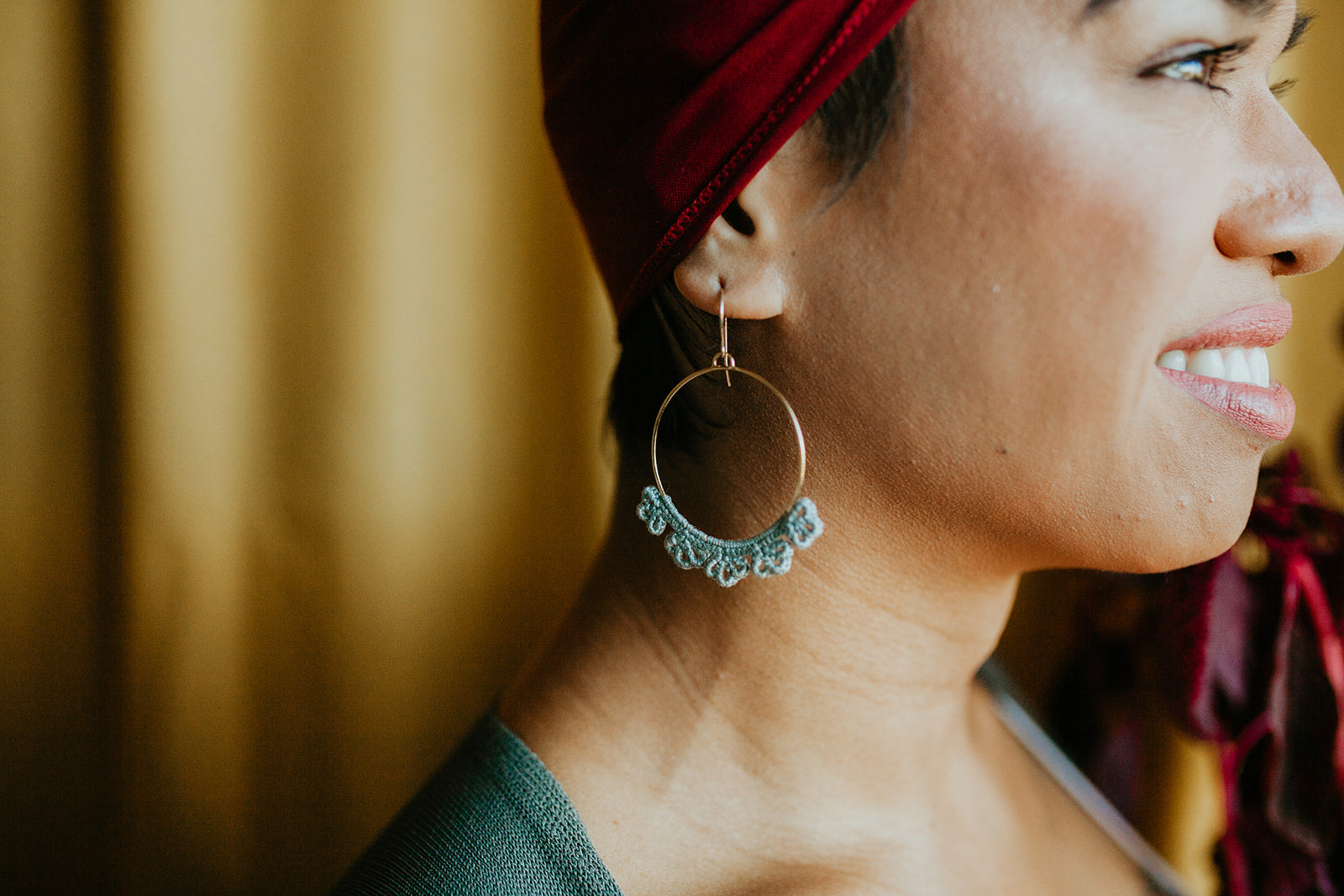 The side profile of a person smiling and wearing a Twyla Dill Large Fleur Earring that is 14kt Gold-Plated and has Slate hand-crocheted lace
