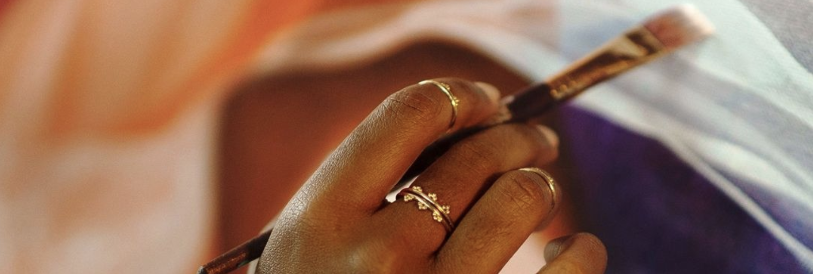 Hand with multiple Twyla Dill Chori Rings on the fingers holding a paintbrush and making a stroke on a painting in the background