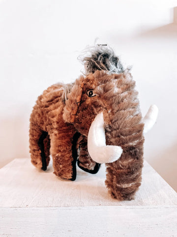 A fuzzy, wooly mammoth dog toy stands on a white background. It is brown with longer gray hair on the top of its head and thick, black stitching around its body.