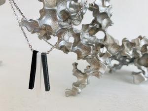 Shown on a white background with an aluminum sculpture, there is a necklace with 3 3D printed pendants. The pendants are long and thin, in silver, white, and black. When turned to the side, names are visible on the pendants.