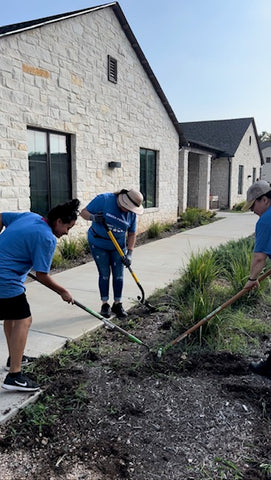 3 women work together gardening. They hold rakes, and move the mulch on the ground. They are volunteering and wear their blue volunteering t shirts