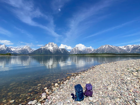 2 backpacks set on pebble beach with view of Teton mountains.