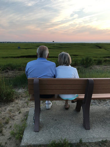View from behind of older couple sitting on bench enjoying sunset over green marsh and water at Greys Beach on Cape Cod